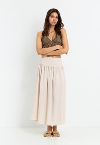 Beige maxi skirt with an elastic waistband, featuring a flowing design. Paired with a brown embroidered cropped top and flat sandals.