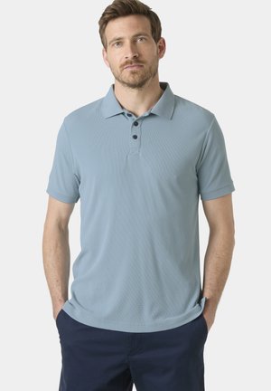 Hombre con barba que lleva una camiseta tipo polo de manga corta de color azul claro y pantalones azul marino, de pie con las manos en los bolsillos frente a un fondo liso.
