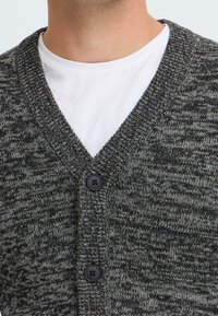 Nærbilde av en person som har på seg en hvit t-skjorte med rund hals under en knappedark grå strikket cardigan.