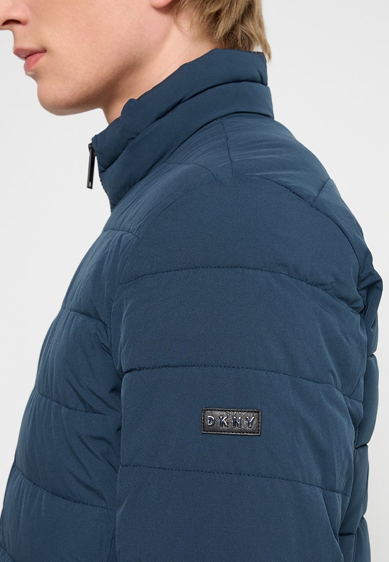 Gequiltede donkerblauwe pufferjas met een hoge kraag, voorzien van een zwart DKNY-logo patch op de mouw en een gladde, zachte textuur.
