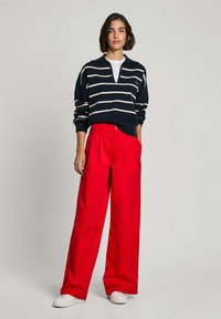 Maglione a righe blu scuro con scollatura a V e tasca frontale abbinato a pantaloni larghi rossi e sneakers bianche, creando un outfit audace e contrastante.