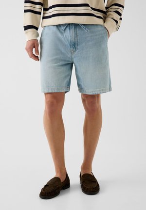 Homme portant un short en jean bleu clair, un pull beige à rayures noires et des mocassins marron foncé, debout les mains dans les poches.