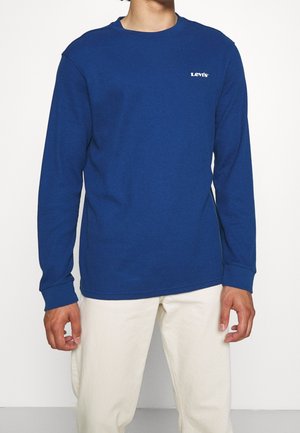 Chemise longue à manches bleues avec une texture côtelée, présentant un petit logo blanc sur la poitrine. Associée à un pantalon de couleur claire.