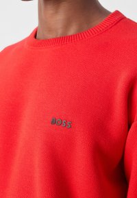 Gros plan sur une personne portant un pull rouge texturé avec un petit logo noir "BOSS" sur la poitrine.