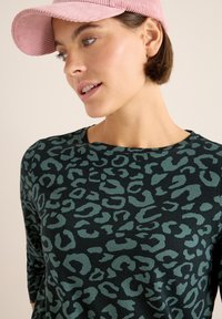 Schwarzes Langarmshirt mit grünem Leopardenmuster, kombiniert mit einer hellrosa Cordmütze. Glattes Material mit Rundhalsausschnitt.
