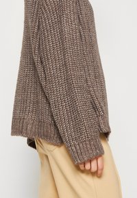 Maglione lavorato a maglia marrone con un motivo testurizzato e polsini a coste, abbinato a pantaloni chiari, che mettono in mostra un fit rilassato.
