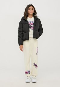 Giacca a vento nera con cappuccio, felpa bianca con grafica e pantaloni della tuta crema con design testuali colorati. Sneakers bianche completano il look.