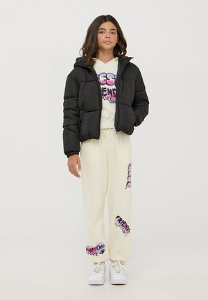 Giacca a vento nera con cappuccio, felpa bianca con grafica e pantaloni della tuta crema con design testuali colorati. Sneakers bianche completano il look.
