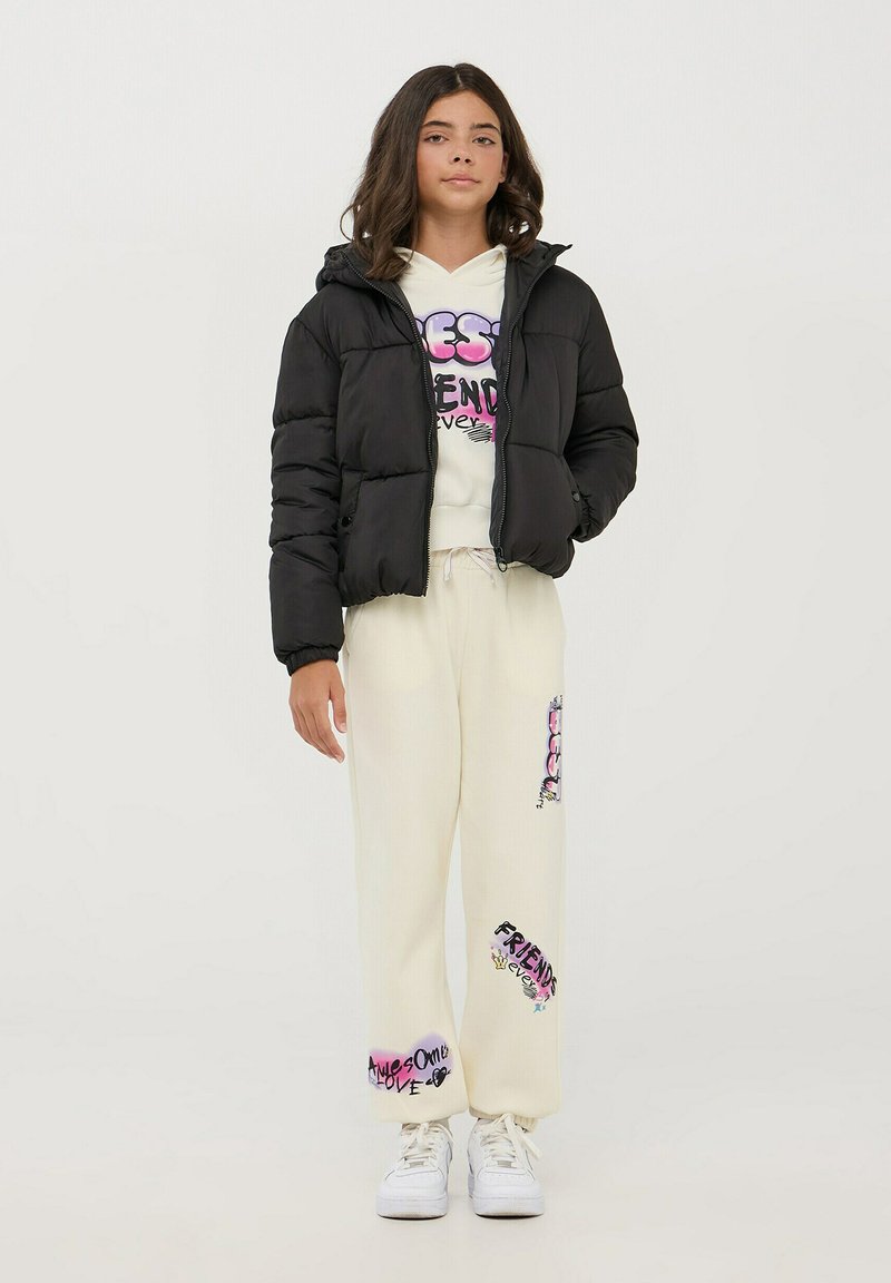 Giacca a vento nera con cappuccio, felpa bianca con grafica e pantaloni della tuta crema con design testuali colorati. Sneakers bianche completano il look.