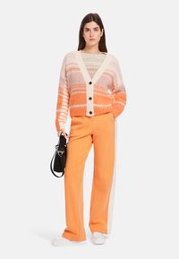 Strickjacke mit orangefarbenen, pinkfarbenen und weißen Streifen, mit V-Ausschnitt und drei Knöpfen. Kombiniert mit orangefarbenen, weiten Hosen und weißen Schuhen.