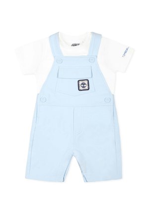 Salopette azzurra realizzata in cotone morbido, con una camicia bianca a maniche corte sotto, un taschino frontale e una toppa con logo.