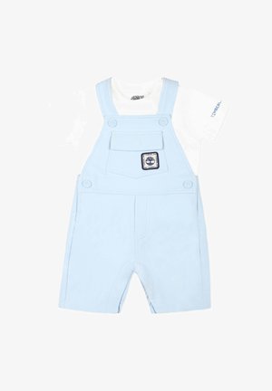 Salopette azzurra realizzata in cotone morbido, con una camicia bianca a maniche corte sotto, un taschino frontale e una toppa con logo.