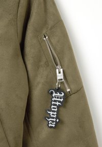 Manga de chaqueta de ante verde oliva con una cremallera plateada y una etiqueta de llavero negra y ornamentada que dice "Utopian." Textura suave.