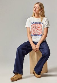 Bijela majica s grafikom sardine i plavim tekstom, uparena s tamnoplavim trapericama širokih nogavica i braon gležnjačama, sjedi na drvenom bloku.