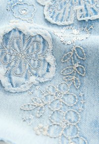 Ljusblå denimtyg med detaljerad vit och beige blommig broderi, fransade kanter och metalltrådsaccenter.