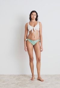 Hvit bikini-topp med knyting foran, kombinert med grønne mønstrede bikini-bunn med sideknyting. Glatt, myk stoff; enkelt design.