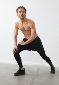 Crne atletske kratke hlače slojevite preko crnih kompresijskih tajica, nosene s crnim tenisicama. Glatka tekstura, prilagodljiv dizajn, minimalno vidljivo brendiranje.