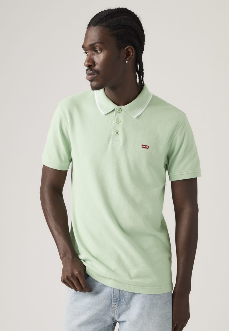 Mann mit geflochtenem Haar, der ein hellgrünes Levi's Poloshirt mit weiß abgesetztem Kragen und hellblaue Jeans trägt und nach links gegen einen schlichten Hintergrund schaut.