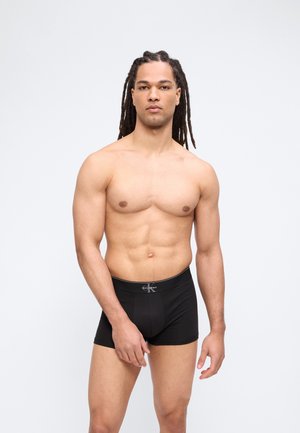 Μυώδης άνδρας με μακριά πλεγμένα μαλλιά φοράει μαύρα εσώρουχα τύπου μποξέρ Calvin Klein, στέκεται μπροστά σε ένα απλό φωτεινό φόντο.