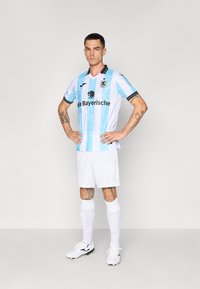 Šviesiai mėlynas ir baltas raištinis futbolininkų marškinėlis su juodu apykakle, turintis juodą logotipą ir tekstą. Pora su baltomis šortais ir kojinėmis.