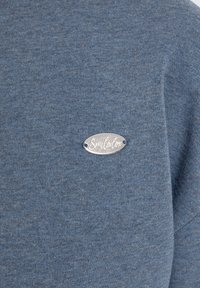 Tessuto di felpa blu con una superficie liscia, dotato di un'etichetta ovale argento con la scritta "Smilodtox" incisa. Design semplice senza ulteriori motivi.