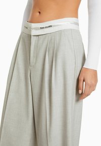 Light gray tailored wide-leg pants with pleats, featuring a satin white waistband inscribed with "TOO CLOSE." Smooth fabric texture.