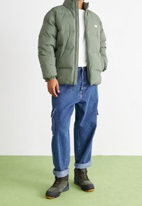 Person i grön puffjacka, vit skjorta, lösa blå cargoj jeans med uppvik, och gröna vandringsskor står på en ljusgrön golv.