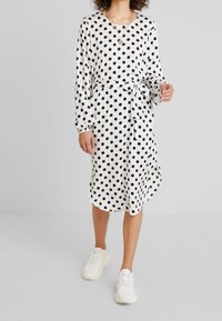 Schwarzes und weißes gepunktetes Kleid mit langen Ärmeln, einem Bindeband in der Taille und einem knielangen Saum. Trägt weiße Sneaker.