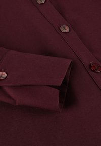 Burgundy button-up shirt with a pointed collar, featuring round brown buttons and a textured fabric. Folded cuff visible on the left side.