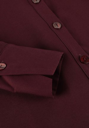 Bordeaux knoopshirt met een puntige kraag, voorzien van ronde bruine knopen en een gestructureerde stof. Opgevouwen manchet zichtbaar aan de linkerkant.