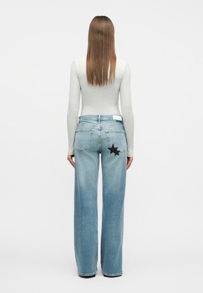 Γυναίκα με ίσια καστανά μαλλιά που φοράει ανοιχτό γκρι μακρυμάνικη μπλούζα και ανοιχτά μπλε φαρδιά τζιν παντελόνια με μαύρο αστέρι στο δεξί τσεπάκι.