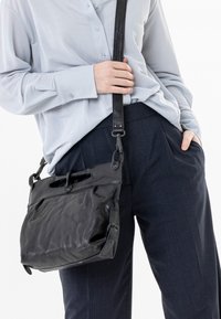 Schwarze Lederhandtasche mit strukturiertem Finish, ausgestattet mit einem oberen Griff und einem verstellbaren Riemen, getragen zu einem hellblauen Hemd und marineblauen Hosen.