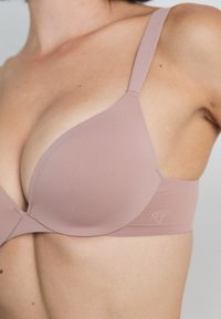 Podstavljeni grudnjak od mekog mauve materijala s podesivim trakama i glatkim čašama. Ima minimalistički logo na pojasu, a detalji šivanja su vidljivi.