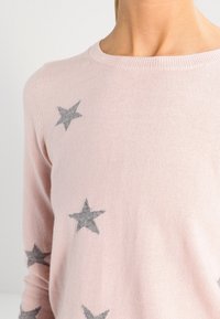 Pull rose pâle à manches longues avec des motifs d'étoiles grises porté par une personne, montrant le cou et le haut du torse sur un fond uni.