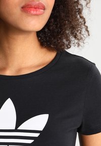 Czarny bawełniany t-shirt z białym graficznym wzorem trójlistnej logo z poziomymi paskami u dołu. Okrągły dekolt, krótki rękaw.