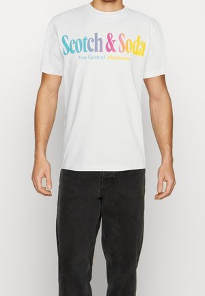 Mężczyzna w białej koszulce z kolorowym tekstem "Scotch & Soda Free Spirit of Amsterdam" i czarnych dżinsach stoi na jednolitym tle.