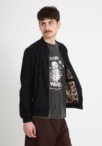 Homme avec moustache et boucles d'oreilles portant une veste noire ouverte montrant une doublure à imprimé léopard, un t-shirt graphique sombre et un pantalon marron, regardant sur le côté.