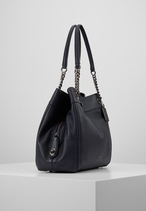 Czarna skórzana torebka na łańcuszku z podwójnymi skórzanymi paskami, przednia kieszeń na zamek oraz metalowa metka z logo, prezentowana na białym piedestale.