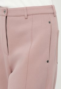 Pantalon rose clair en tissu doux ; présente un devant plat, des poches latérales et un petit détail en bouton noir sur la taille.