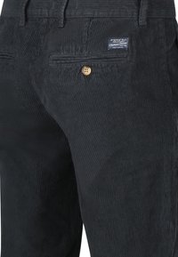 Donkergrijze corduroy broek met een achterzak met knop en een klein blauw stoffen merklabel boven de zak.