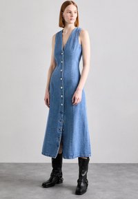 Denim-kjole med dyp V-hals og knappelukking. A-linje silhuett, kne-lengde, i lyseblå med kontrasterende hvite knapper, parret med svarte støvler.