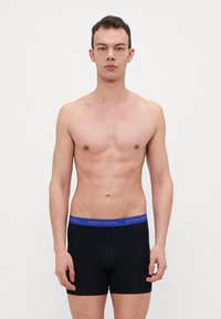 Boxers noirs avec une ceinture élastique bleue portant le texte "BJÖRN BORG." Tissu lisse, design ajusté et longueur jusqu'au milieu de la cuisse.