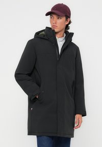 Zwarte waterdichte parka met een capuchon, met een ritssluiting aan de voorkant, zijzakken en een voering aan de binnenkant. Draag het over een gestreept shirt en blauwe jeans.
