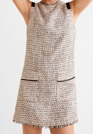 Mouwloze, getextureerde beige en zwarte tweedjurk met twee voorzakken en een rafelige zoom, gedragen door een persoon met opgeheven armen.