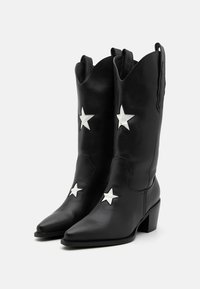 Czarne skórzane kowbojskie buty z wydłużonymi czubkami, z białymi gwiazdami i na podwyższonej podeszwie. Projekt zawiera falbaniasty brzeg na górze.