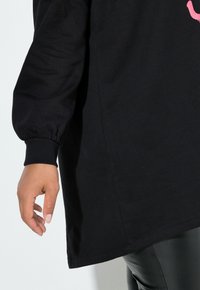 Tunic nera a maniche lunghe con polsini elasticizzati e orlo arrotondato, con una mano appoggiata sul fianco, disposta su uno sfondo chiaro.