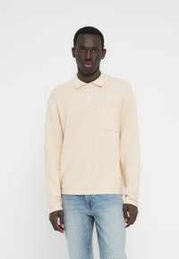 Πόλο μπλούζα από υφασμάτινη μπεζ υφή με πατιλέτα τριών κουμπιών και τσέπη εφαρμογής. Συνδυασμένη με ανοιχτόχρωμο τζιν παντελόνι.