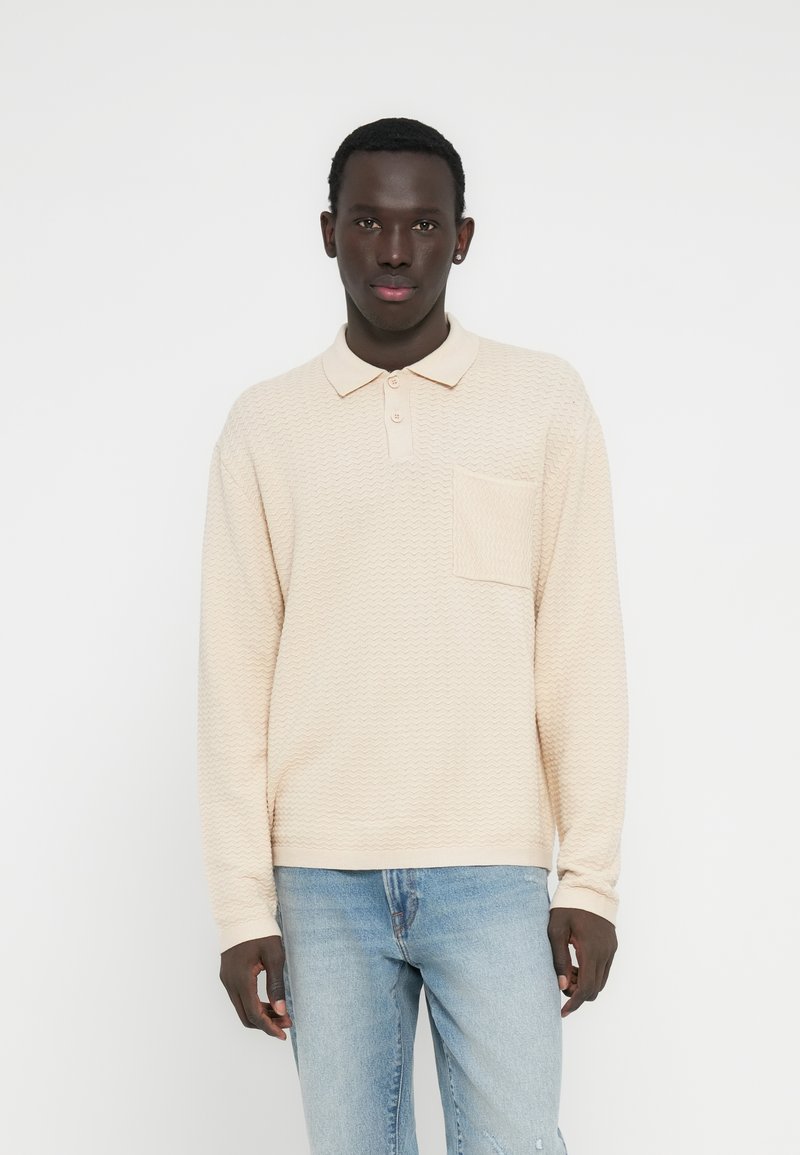 Πόλο μπλούζα από υφασμάτινη μπεζ υφή με πατιλέτα τριών κουμπιών και τσέπη εφαρμογής. Συνδυασμένη με ανοιχτόχρωμο τζιν παντελόνι.