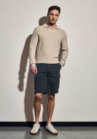 Beige pletený svetr s výstřihem do V a dlouhými rukávy, doplněný tmavými cargo šortkami s bočními kapsami a bílými teniskami s béžovými akcenty.