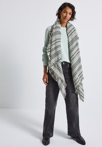 Lys grøn sweater, sorte vide jeans og et stort sort-hvidt mønstret tørklæde med frynser. Enkel design og blød tekstur.